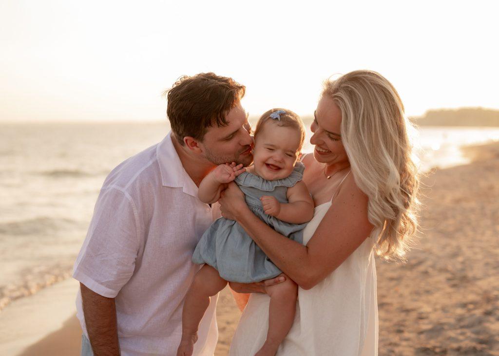 Mother and father holding their baby girl, laughing together on the beach at sunset during a family photography session.